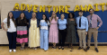 Eight adults smiling in front of gold "ADVENTURE AWAITS" balloons.
